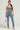 A person stands confidently in the JOLIE rhinestone-embellished denim corset paired with matching jeans featuring distressed detailing. They hold a glittery silver handbag and wear silver high heels, accessorized with large dangling earrings, against a plain light gray background.
