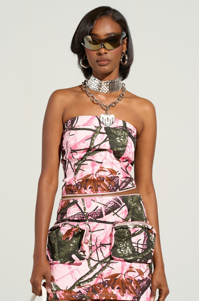 A person confidently stands wearing the versatile WEAR IT TWO WAYS TOP OR DRESS in a strapless pink camouflage design, paired with a matching skirt featuring large pockets. Their dark hair is complemented by stylish sunglasses and bold silver jewelry, including a chunky necklace. The ensemble seamlessly transforms into a chic convertible dress against the plain background.