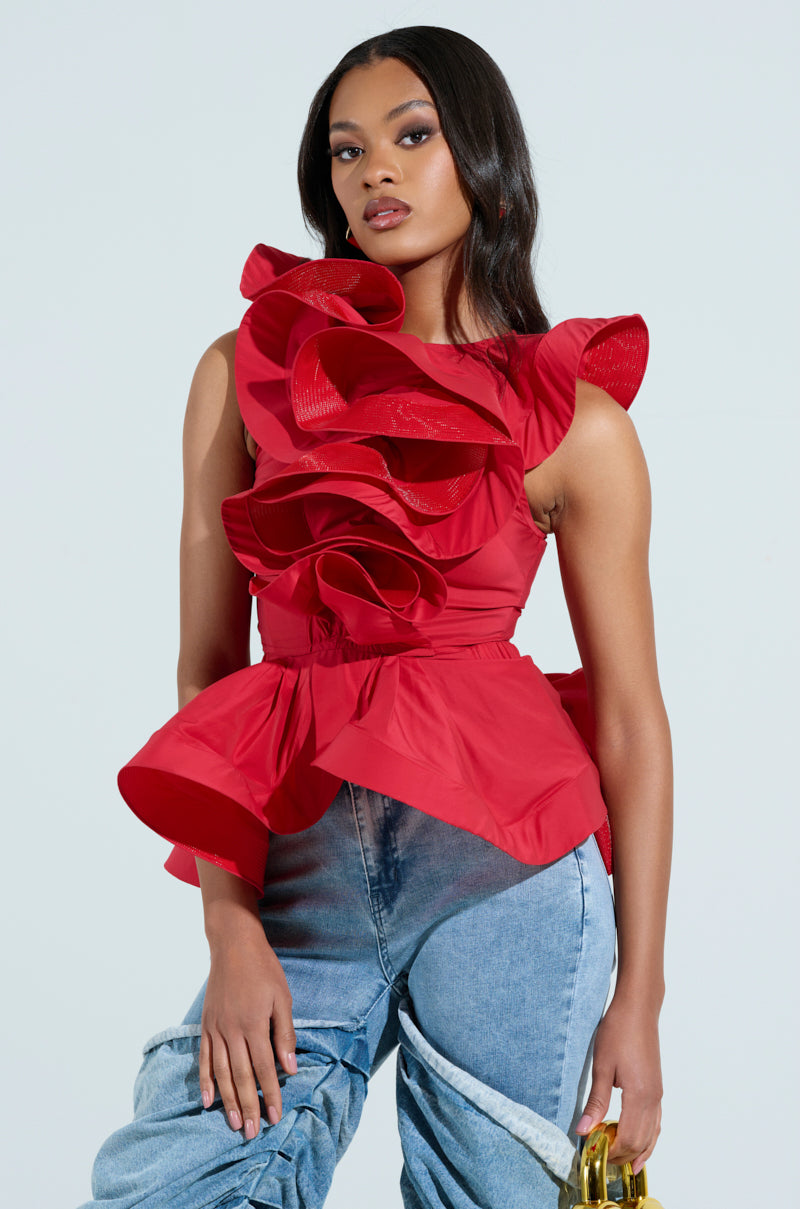 A person confidently stands against a light background in the MISS FASHIONISTA Sleeveless Ruffle Peplum Blouse in vibrant red, paired with blue jeans and holding a small yellow handbag. Their dark hair cascades over their shoulders.