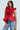 A person confidently stands against a light background in the MISS FASHIONISTA Sleeveless Ruffle Peplum Blouse in vibrant red, paired with blue jeans and holding a small yellow handbag. Their dark hair cascades over their shoulders.