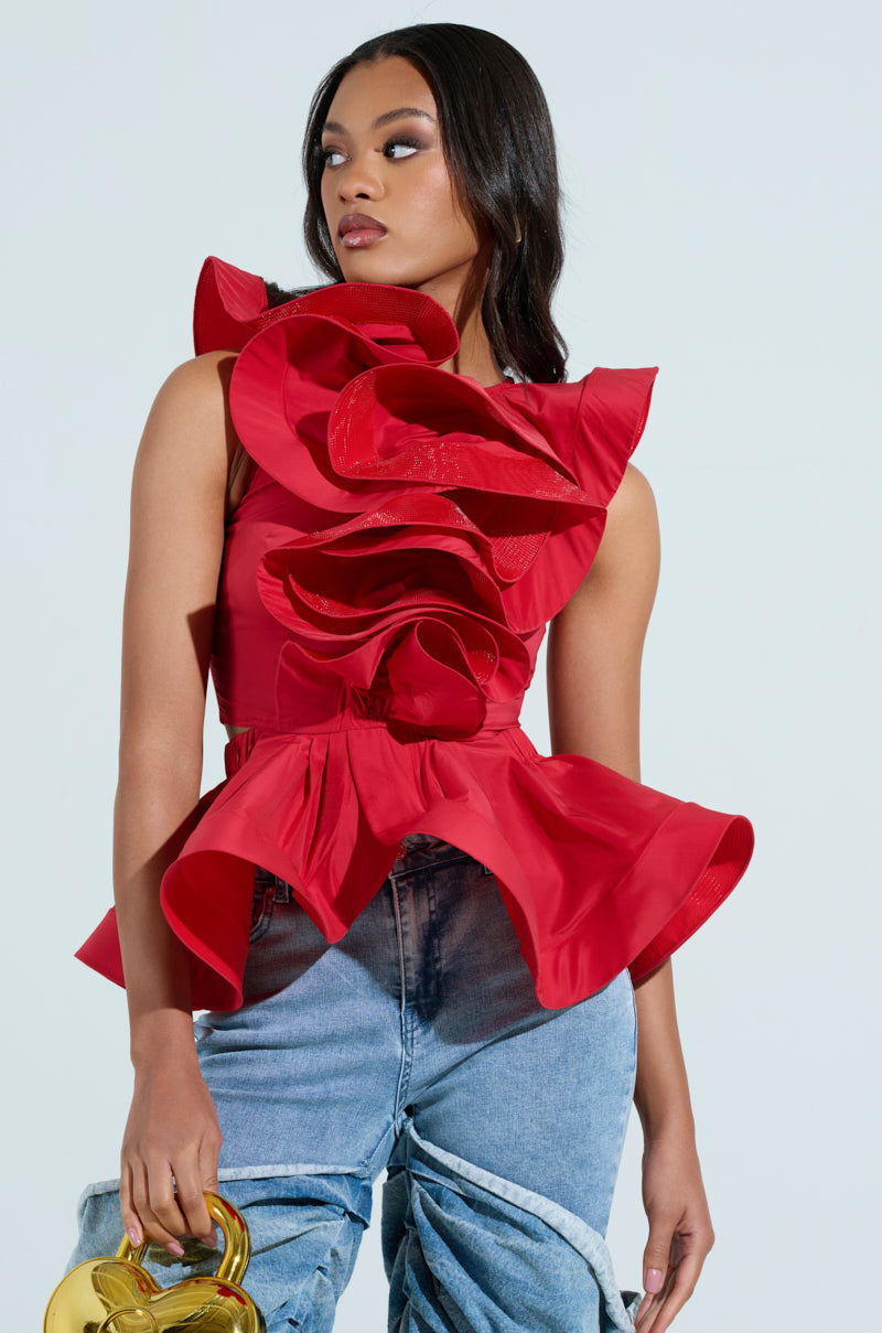 A woman confidently poses in a MISS FASHIONISTA sleeveless red ruffle peplum blouse, holding a gold sculptural handbag and wearing light blue distressed jeans against a plain light background, highlighting her stylish look.