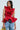 A woman confidently poses in a MISS FASHIONISTA sleeveless red ruffle peplum blouse, holding a gold sculptural handbag and wearing light blue distressed jeans against a plain light background, highlighting her stylish look.