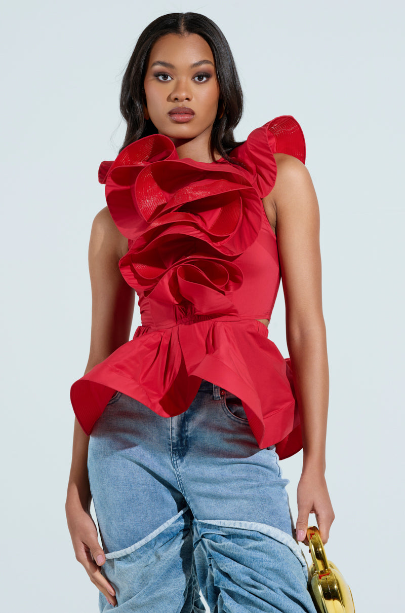 Against a light background, someone wears the MISS FASHIONISTA sleeveless ruffle peplum blouse in red, crafted from a cotton blend, paired with light blue jeans featuring unique pleating. They hold a gold purse. The blouse showcases dramatic floral-like fabric details.