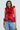 Against a light background, someone wears the MISS FASHIONISTA sleeveless ruffle peplum blouse in red, crafted from a cotton blend, paired with light blue jeans featuring unique pleating. They hold a gold purse. The blouse showcases dramatic floral-like fabric details.