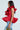 Wearing the MISS FASHIONISTA Sleeveless Ruffle Peplum Blouse in vibrant red, a long-haired individual pairs it with light blue jeans and holds a small gold handbag, casting a sideways glance against a plain background.