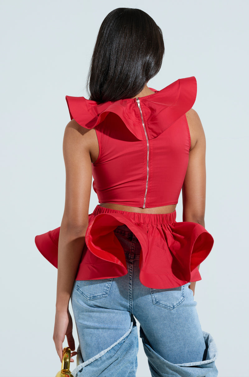 A woman with long hair wears the Miss Fashionista Sleeveless Ruffle Peplum Blouse in red and light blue jeans. Facing away, she holds a gold object as a denim jacket drapes over one arm against a neutral backdrop.