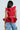 A woman with long hair wears the Miss Fashionista Sleeveless Ruffle Peplum Blouse in red and light blue jeans. Facing away, she holds a gold object as a denim jacket drapes over one arm against a neutral backdrop.
