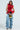 A woman poses against a plain backdrop wearing the MISS FASHIONISTA Sleeveless Ruffle Peplum Blouse in red with loose denim pants, holding a heart-shaped gold object. She completes her look with leopard-print shoes, exuding a composed and confident demeanor.