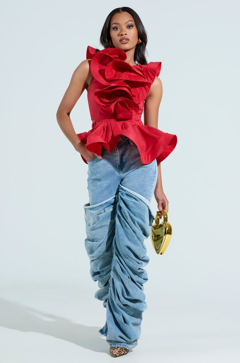 The individual sports a MISS FASHIONISTA Sleeveless Ruffle Peplum Blouse in vibrant red paired with unique ruched denim pants. They accessorize with a heart-shaped gold handbag and leopard print shoes against a plain white backdrop.