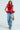 The individual sports a MISS FASHIONISTA Sleeveless Ruffle Peplum Blouse in vibrant red paired with unique ruched denim pants. They accessorize with a heart-shaped gold handbag and leopard print shoes against a plain white backdrop.