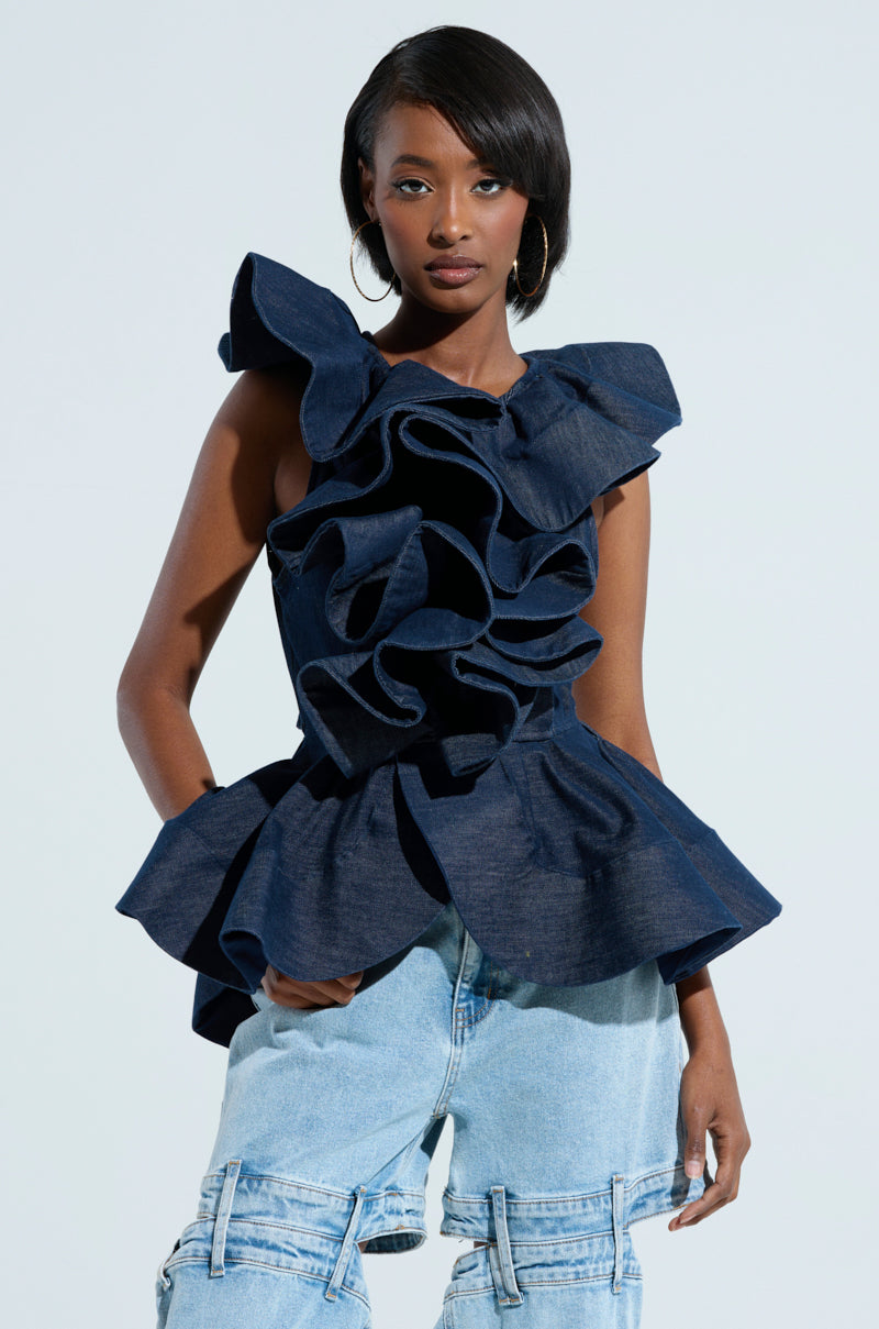 A person stands confidently wearing the MISS FASHIONISTA SLEEVELESS BLOUSE, a dark blue peplum silhouette with ruffled detailing, paired with light denim jeans. They sport hoop earrings and short, styled hair, posed against a plain, light background.