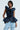 A person stands confidently wearing the MISS FASHIONISTA SLEEVELESS BLOUSE, a dark blue peplum silhouette with ruffled detailing, paired with light denim jeans. They sport hoop earrings and short, styled hair, posed against a plain, light background.