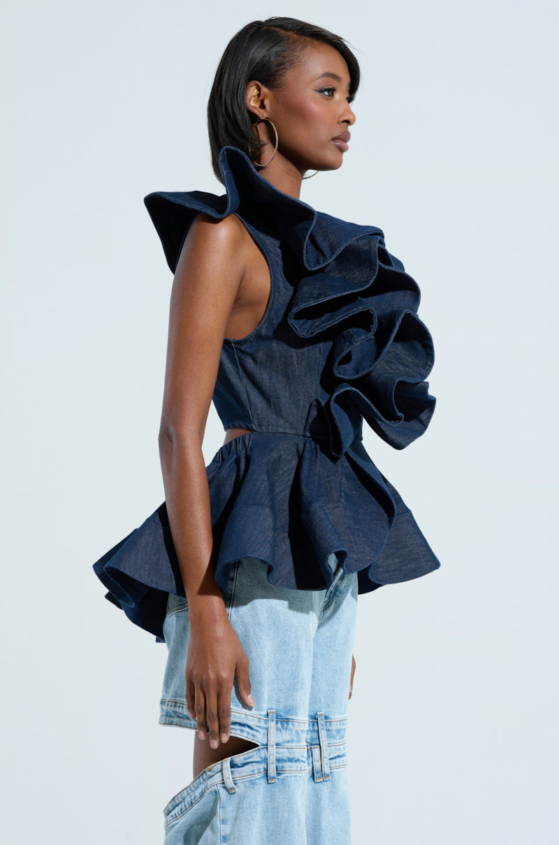 A person wearing a dark blue MISS FASHIONISTA SLEEVELESS BLOUSE by AKIRA Label, featuring dramatic, layered ruffles from shoulder to waist, is paired with two-tone light blue denim pants. The peplum silhouette of the top enhances the unique design of the pants against a plain background.