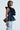 A person wearing a dark blue MISS FASHIONISTA SLEEVELESS BLOUSE by AKIRA Label, featuring dramatic, layered ruffles from shoulder to waist, is paired with two-tone light blue denim pants. The peplum silhouette of the top enhances the unique design of the pants against a plain background.