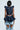 A person with short black hair is facing away, showcasing a MISS FASHIONISTA SLEEVELESS BLOUSE featuring a large ruffle collar and zipper, paired with unique denim shorts that have open panels and ruffled edges. The background is light and neutral.