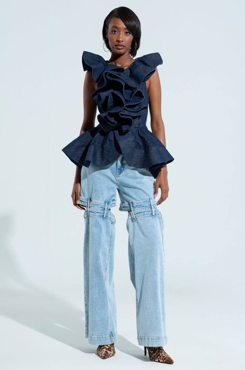 A woman is modeling an AKIRA Label fashion ensemble featuring the MISS FASHIONISTA SLEEVELESS BLOUSE with ruffled details and light blue baggy jeans. She completes the look with leopard print boots, standing out against the plain white background.