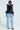 A woman is modeling an AKIRA Label fashion ensemble featuring the MISS FASHIONISTA SLEEVELESS BLOUSE with ruffled details and light blue baggy jeans. She completes the look with leopard print boots, standing out against the plain white background.