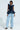 Clad in the chic MISS FASHIONISTA SLEEVELESS BLOUSE, a person stands confidently, teamed with light blue wide-leg jeans accented by multiple belts. The ensemble is elevated by the addition of leopard print heels against a plain white backdrop, highlighting the fashion-forward look.