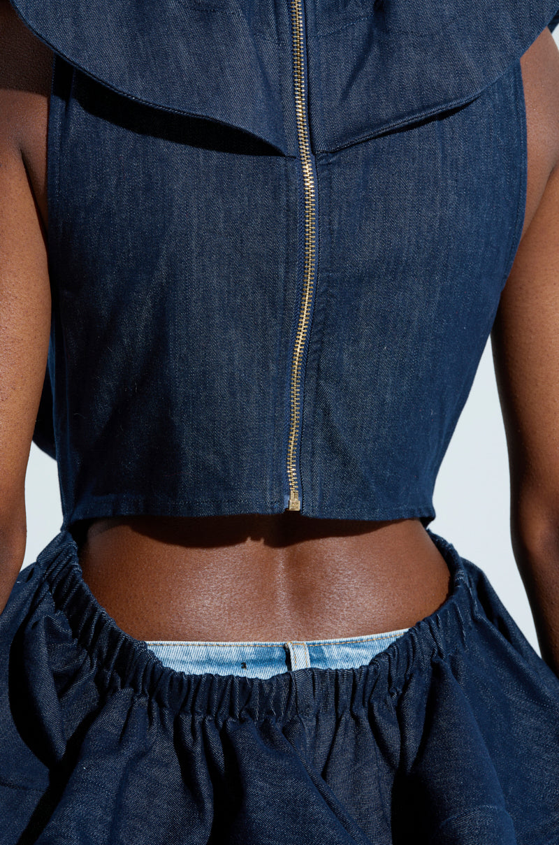 The individual is styled in an AKIRA Label MISS FASHIONISTA SLEEVELESS BLOUSE with a zipper, paired with a ruffled denim skirt. The outfit features a chic cutout detail that reveals parts of the waist and back against a plain, light-colored background.