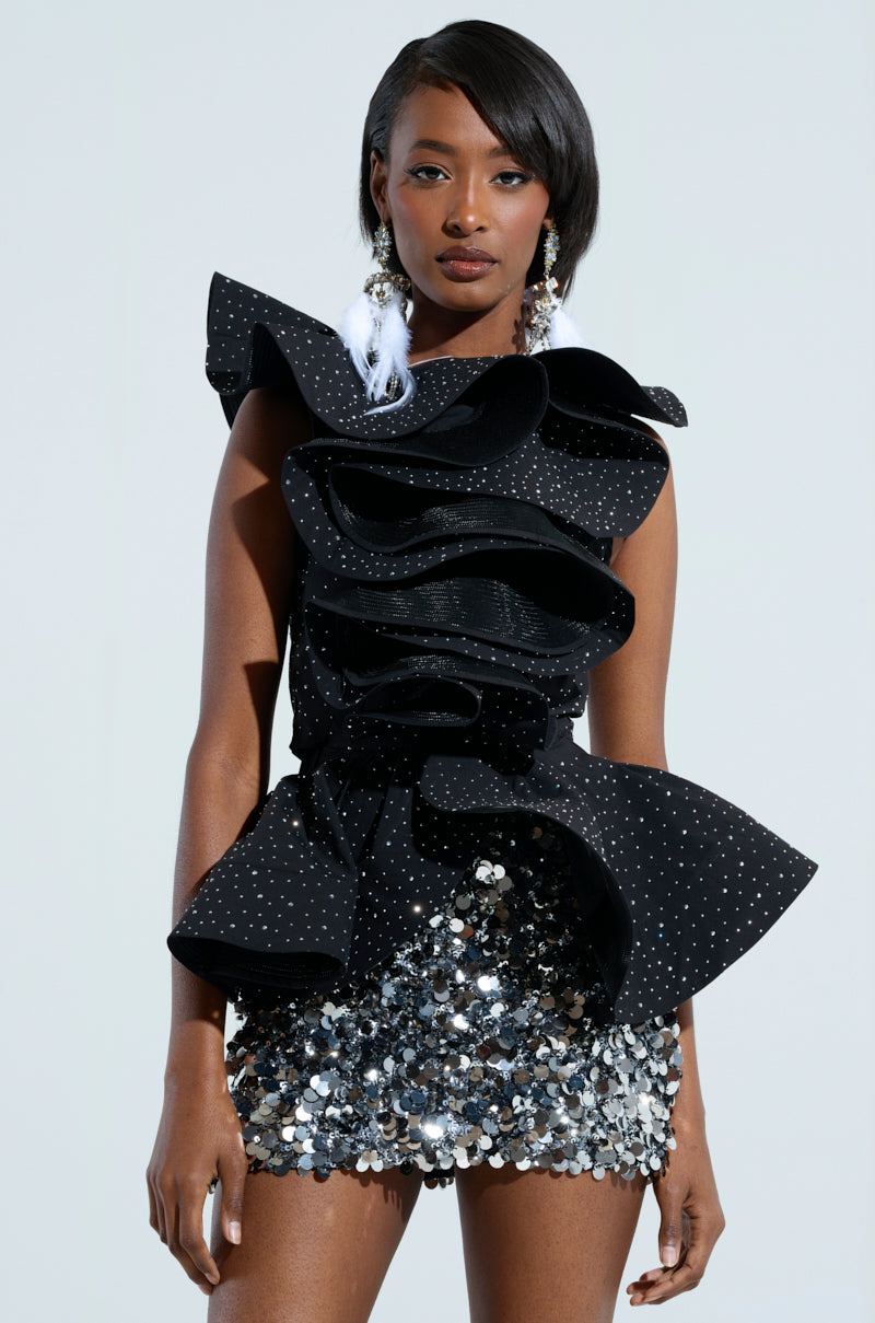 A woman wearing a sleeveless rhinestone blouse, showcasing the MS FASHIONISTA design, paired with a sequined silver mini skirt. Her long, straight hair enhances her look accented by large, decorative earrings, all highlighted beautifully against the light-colored background.