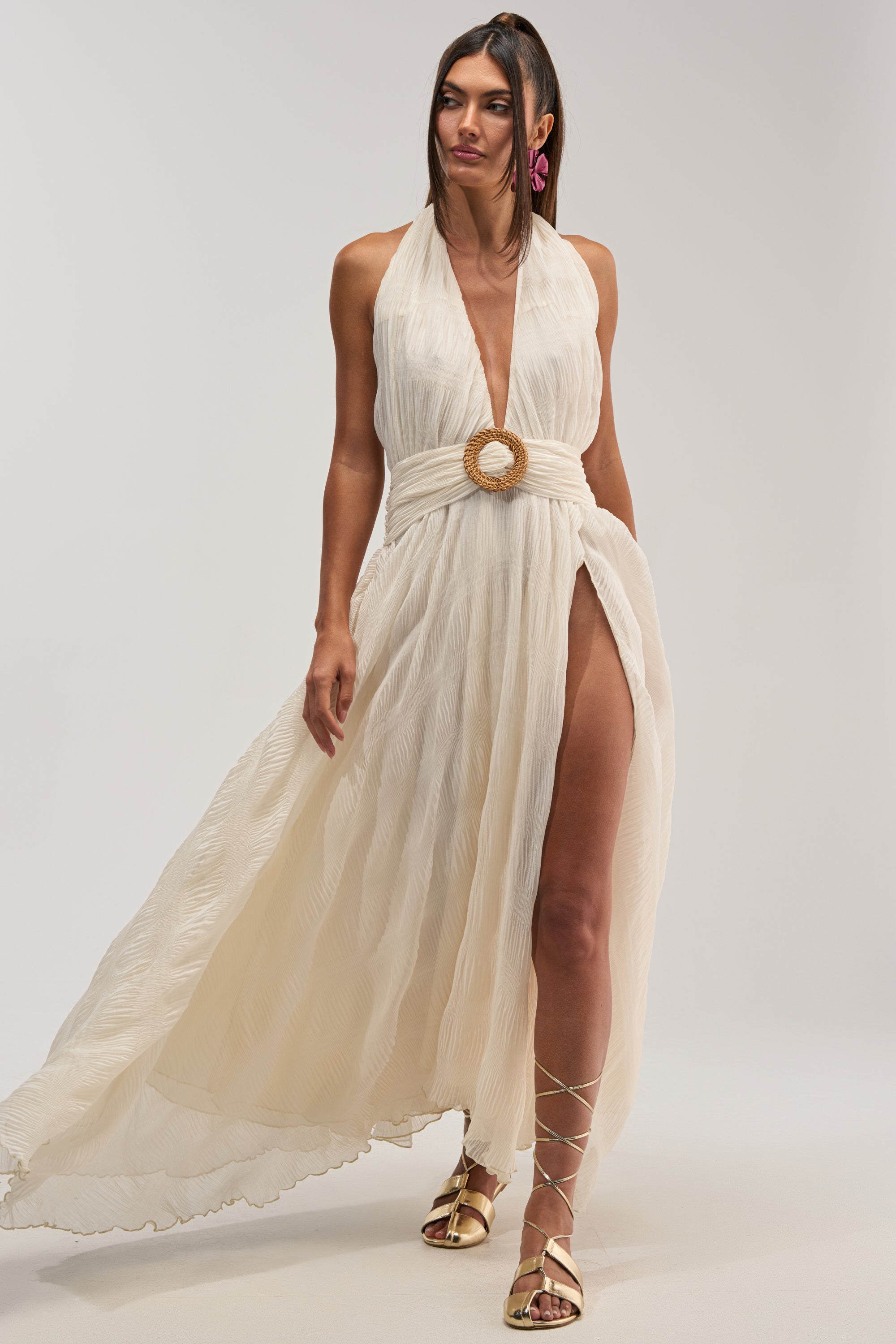 A woman styles the BEACH CLUB BELTED MAXI DRESS IN WHITE—a sleeveless halter maxi with pleated chiffon, deep slit, and bold circular belt buckle—with pink earrings, gold strappy sandals, and loose flowing hair for a chic summer look.