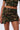 A person wearing the NO MERCY CAMO MINI SKIRT with gold stud accents and rhinestone embellishments. The ensemble features a cropped button-up top paired with a mini skirt that has cargo pockets and front slits on both thighs. The background is plain white.