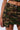 A person is wearing the NO MERCY CAMO MINI SKIRT, which is adorned with rhinestone embellishments. The camo-patterned skirt features side pockets, a front button, and a short slit on the left side. The upper part of the body is also partially visible in a matching top.