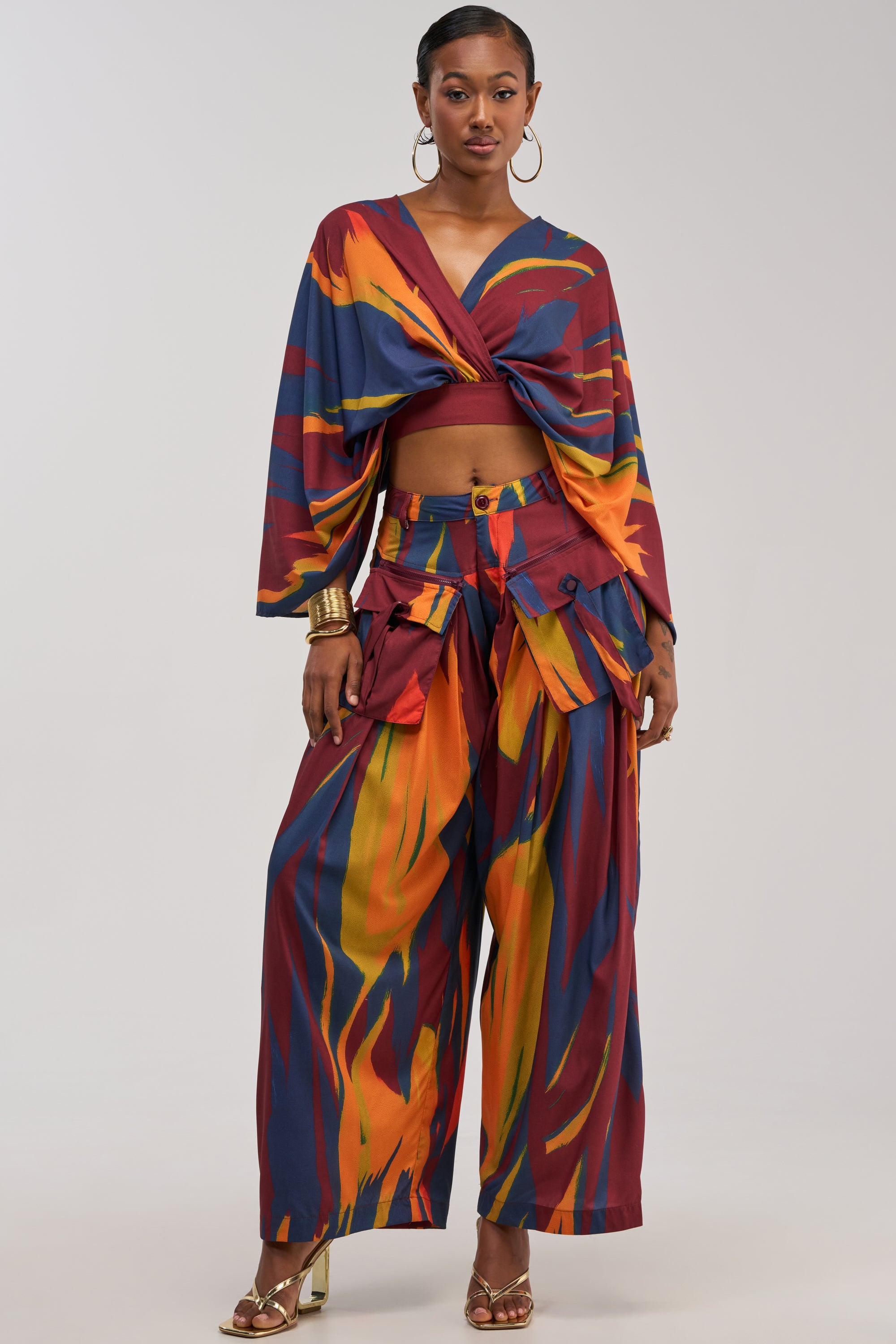 A woman poses in the TAKE ME AWAY TROUSER, featuring a colorful abstract multicolor print and high-rise wide-leg silhouette, paired with a matching twisted crop top and accessorized with hoop earrings, gold bangles, and heeled sandals.
