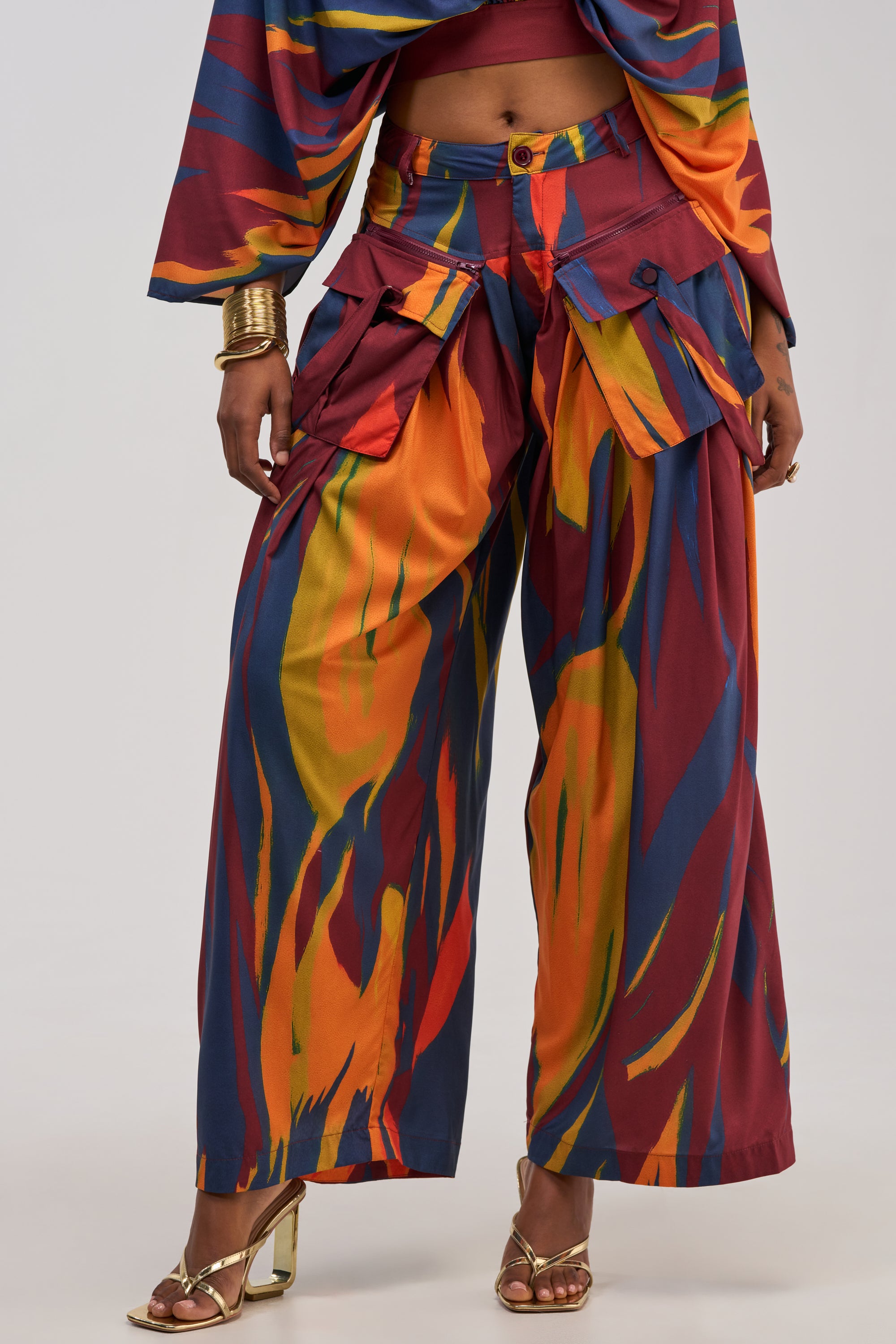 A person wearing the TAKE ME AWAY TROUSER, a pair of wide-leg, high-rise pants with a bold abstract multicolor print in vibrant red, yellow, orange, and blue hues. The trousers feature large front pockets and are styled with gold heels and chunky gold bracelets. The upper body is partially visible.