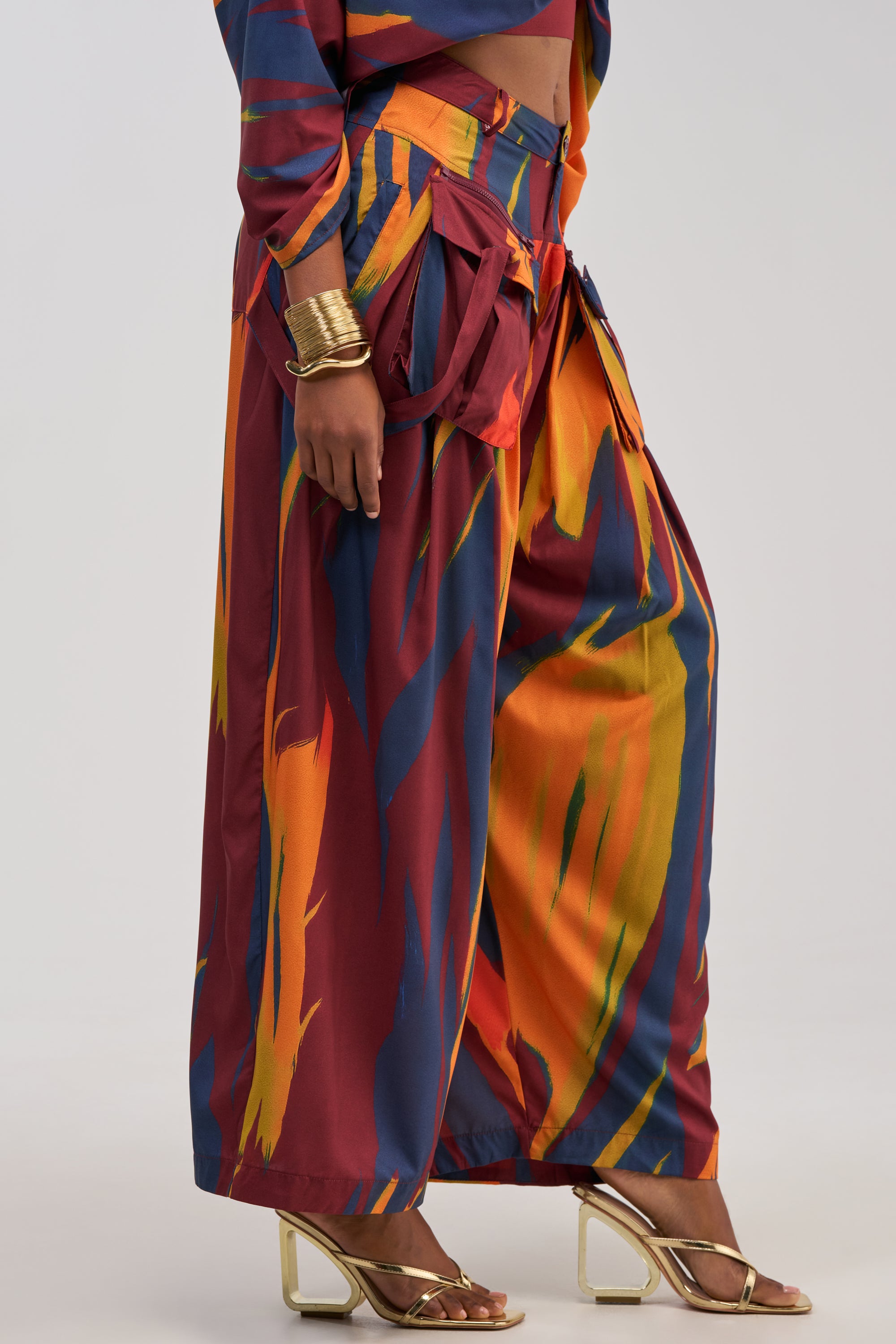 A person wearing the TAKE ME AWAY TROUSER, which features a high-rise, wide-leg silhouette and a striking abstract multicolor print, paired with a coordinating top, gold high-heeled sandals, and several gold bangles on their wrist.