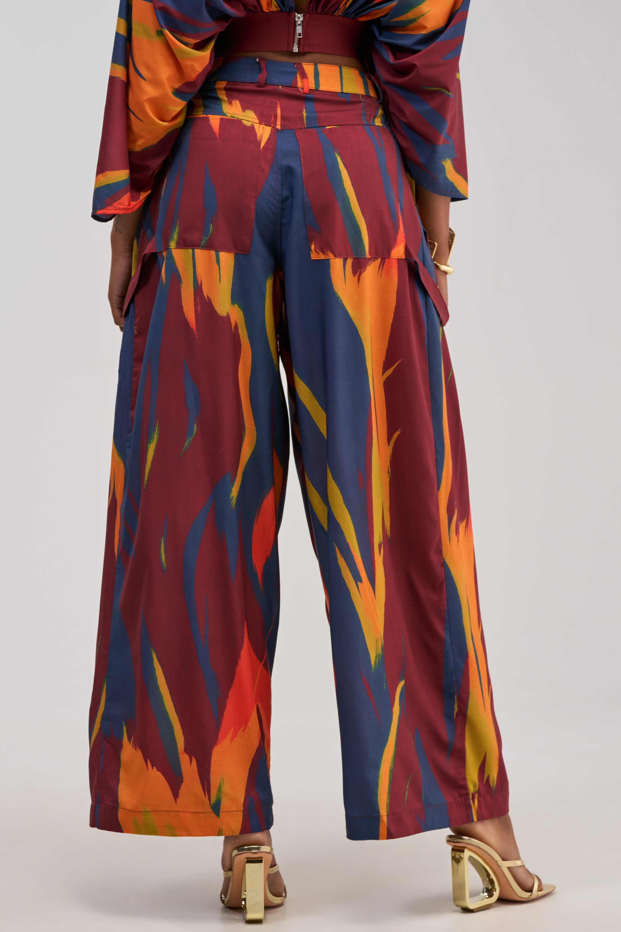 A person wearing the TAKE ME AWAY TROUSER, featuring a high-rise, wide-leg silhouette and a bold abstract multicolor print in red, yellow, orange, and blue. They are standing and wearing heeled sandals.