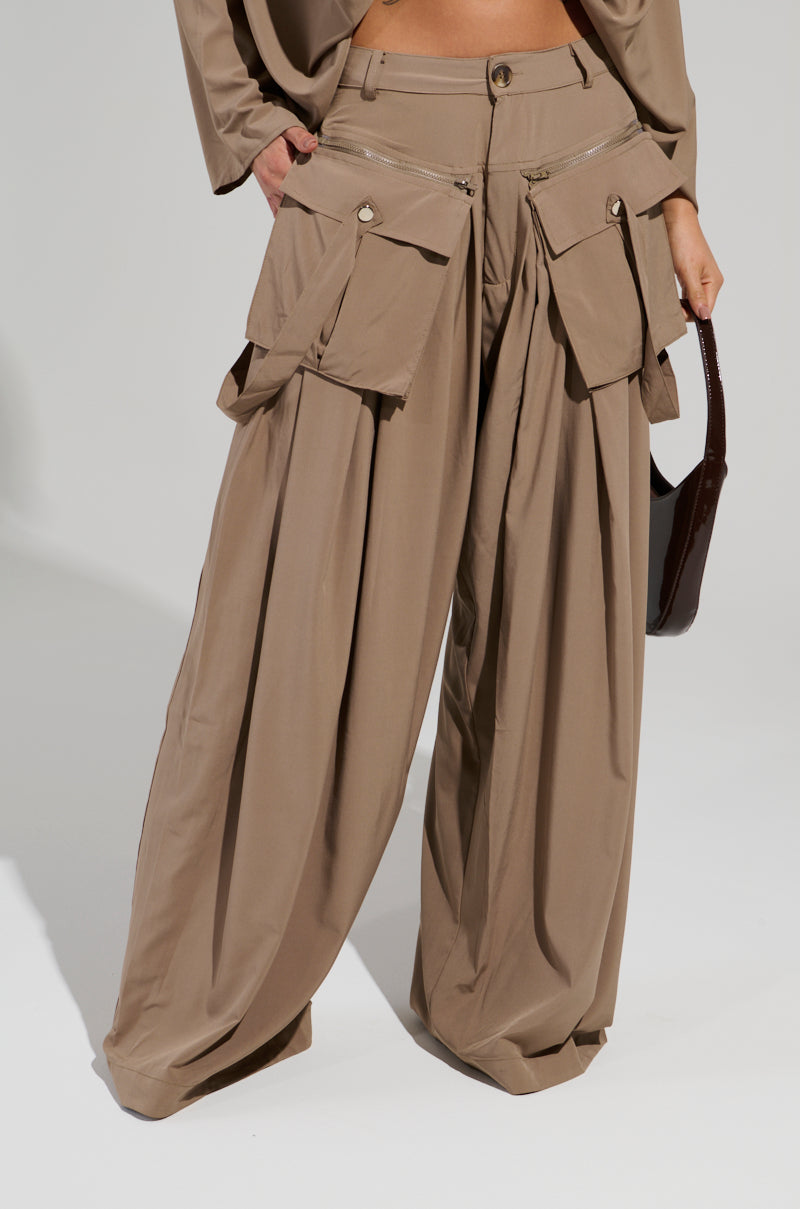A person wearing the AKIRA Label TALK ABOUT ME TROUSER, which showcases a wide-leg silhouette and high-rise design, complete with several spacious pockets. The outfit is paired with a matching top, and the individual carries a dark handbag against a simple, light-colored backdrop.