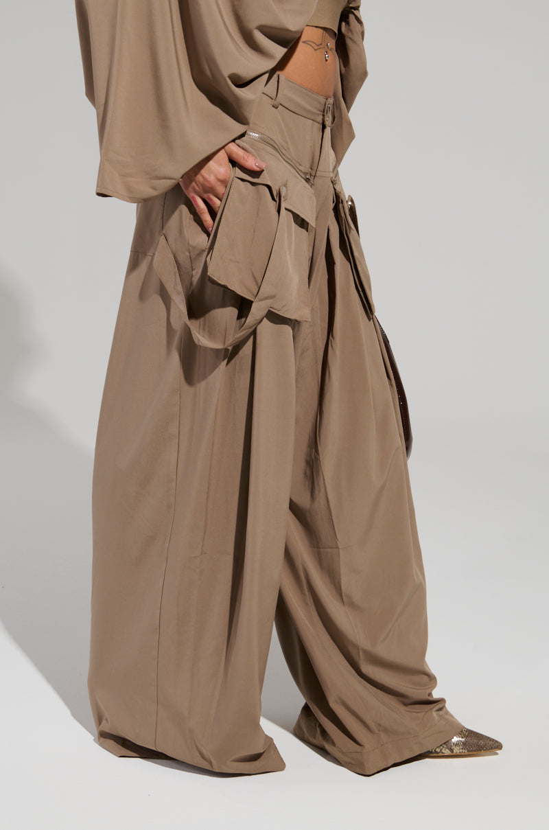 A person dressed in a beige ensemble showcases the AKIRA Label TALK ABOUT ME TROUSER, noted for its wide-leg silhouette and multiple pockets, paired with a loose-fitting top. They complete the look with pointed-toe shoes against a simple, light-colored background.