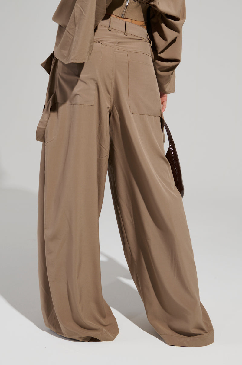 The image depicts a person from the back, dressed in the TALK ABOUT ME TROUSER by AKIRA Label, which boasts a wide leg silhouette and high waist design with large side pockets. They wear a coordinating beige top and hold a dark-colored accessory in one hand.