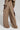 The image depicts a person from the back, dressed in the TALK ABOUT ME TROUSER by AKIRA Label, which boasts a wide leg silhouette and high waist design with large side pockets. They wear a coordinating beige top and hold a dark-colored accessory in one hand.