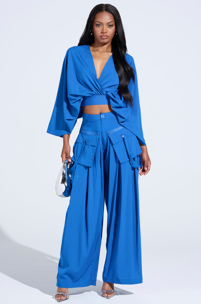 A woman confidently poses in vibrant blue, pairing a knotted crop top with TALK ABOUT ME BLUE TROUSER, featuring cargo pockets. She holds a shiny silver bag and wears silver heels against a light background, her long dark hair styled straight. 0