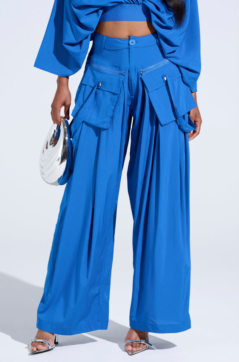 The model is styled in the TALK ABOUT ME BLUE TROUSER, a high-rise wide-leg piece with cargo-style pockets, matched with a coordinating cropped top from the AKIRA Label. They accessorize with a shiny silver handbag and silver heels against a plain backdrop. 1