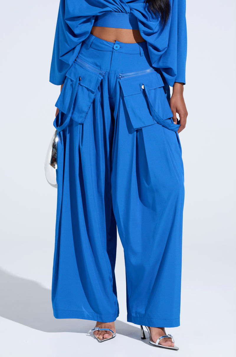 Someone is wearing the vibrant blue TALK ABOUT ME TROUSER, featuring high-rise wide-leg design and cargo-style pockets, with a matching cropped AKIRA Label top. They accessorize with a silver handbag and high-heeled sandals against a stark white backdrop, making the outfit stand out. 2