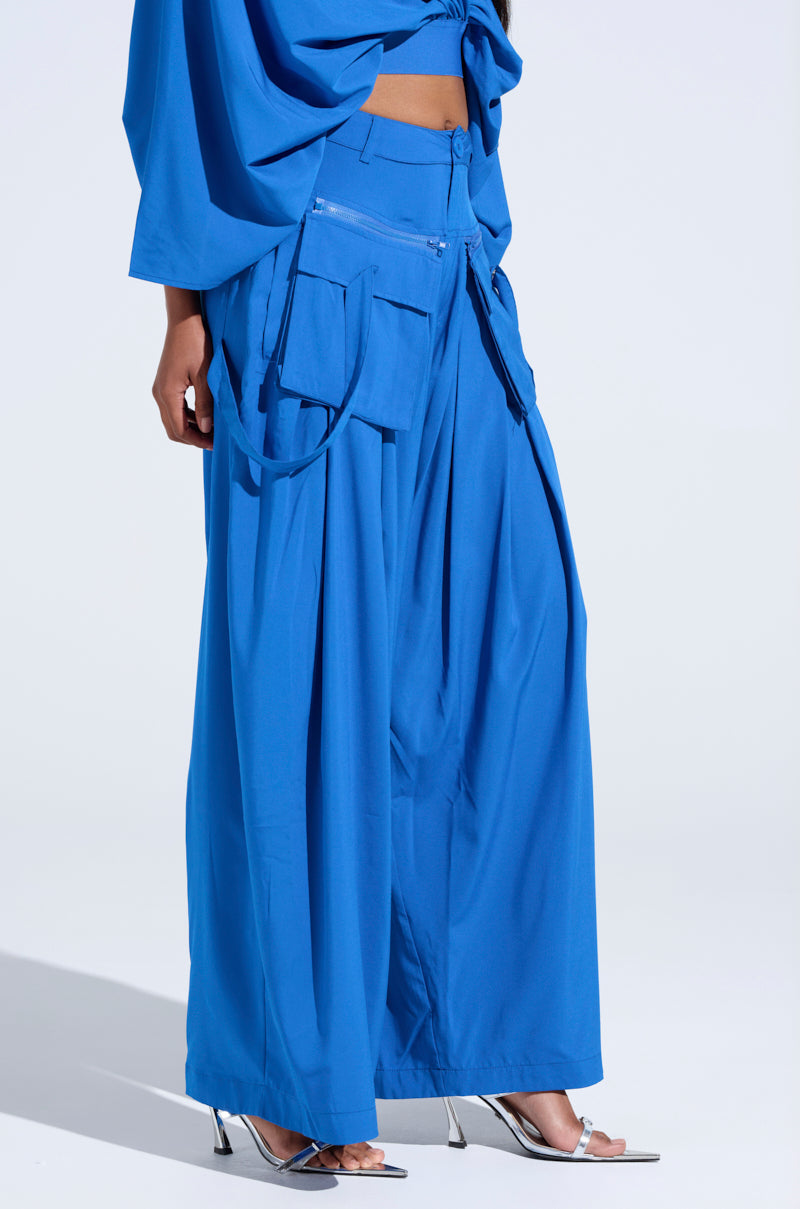A person dons the AKIRA Label's TALK ABOUT ME BLUE TROUSER, featuring bright blue high-rise wide-leg design, matched with a top adorned with large cargo-style pockets and adjustable straps. The ensemble is elegantly paired with silver high-heeled sandals against a light background. 3