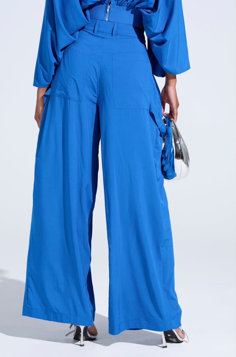 A person elegantly wears the TALK ABOUT ME BLUE TROUSER from the AKIRA Label, featuring bright blue, high-rise wide-leg design with cargo-style pockets and a matching top. They hold a metallic purse and stand gracefully in silver high-heeled shoes against a plain white background. 4