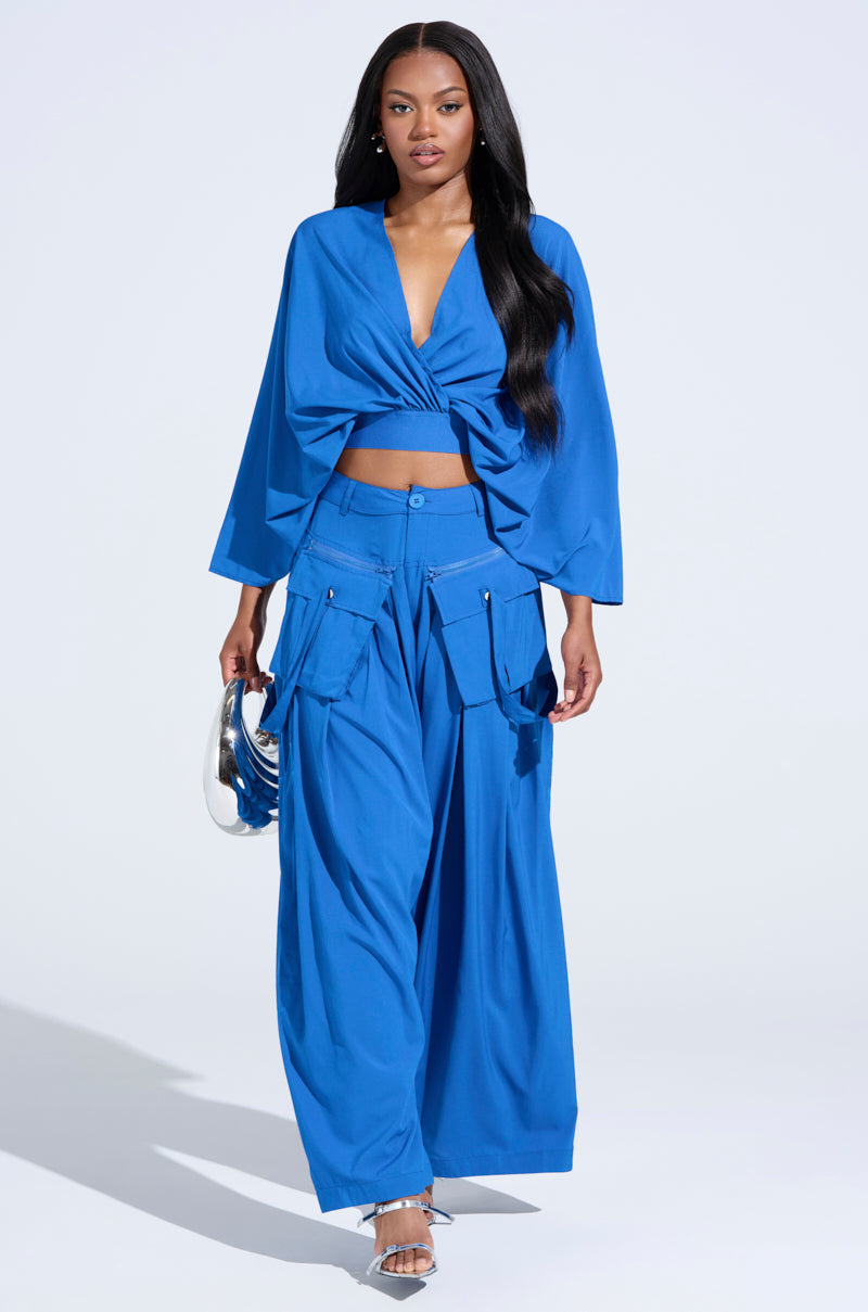 A person wears the vibrant blue TALK ABOUT ME BLUE TROUSER with cargo-style pockets, paired with a long-sleeve crop top. They accessorize with a small silver handbag and transparent heels against a plain white background. 5