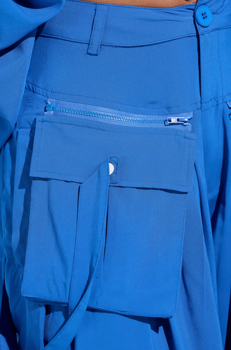 Close-up of TALK ABOUT ME BLUE TROUSER by AKIRA Label showcases vibrant blue high-rise design with a front zipper and cargo-style flap pocket. Smooth fabric and button detail on the waistband enhance its modern utilitarian aesthetic. 6