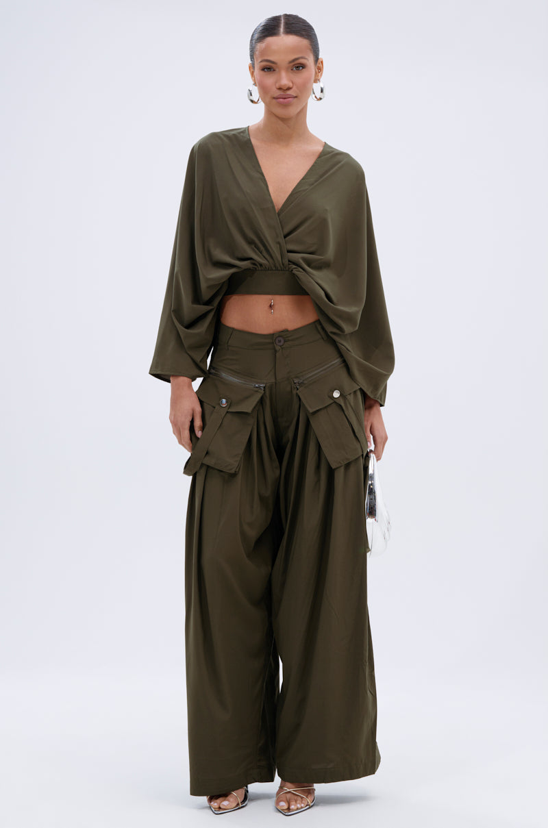 A woman poses against a plain background in olive, wearing the TALK ABOUT ME TROUSER—high-rise, wide-leg trousers with pleats—and a draped, long-sleeve v-neck crop top. She accessorizes with a small white bag, hoop earrings, and sandals. 0