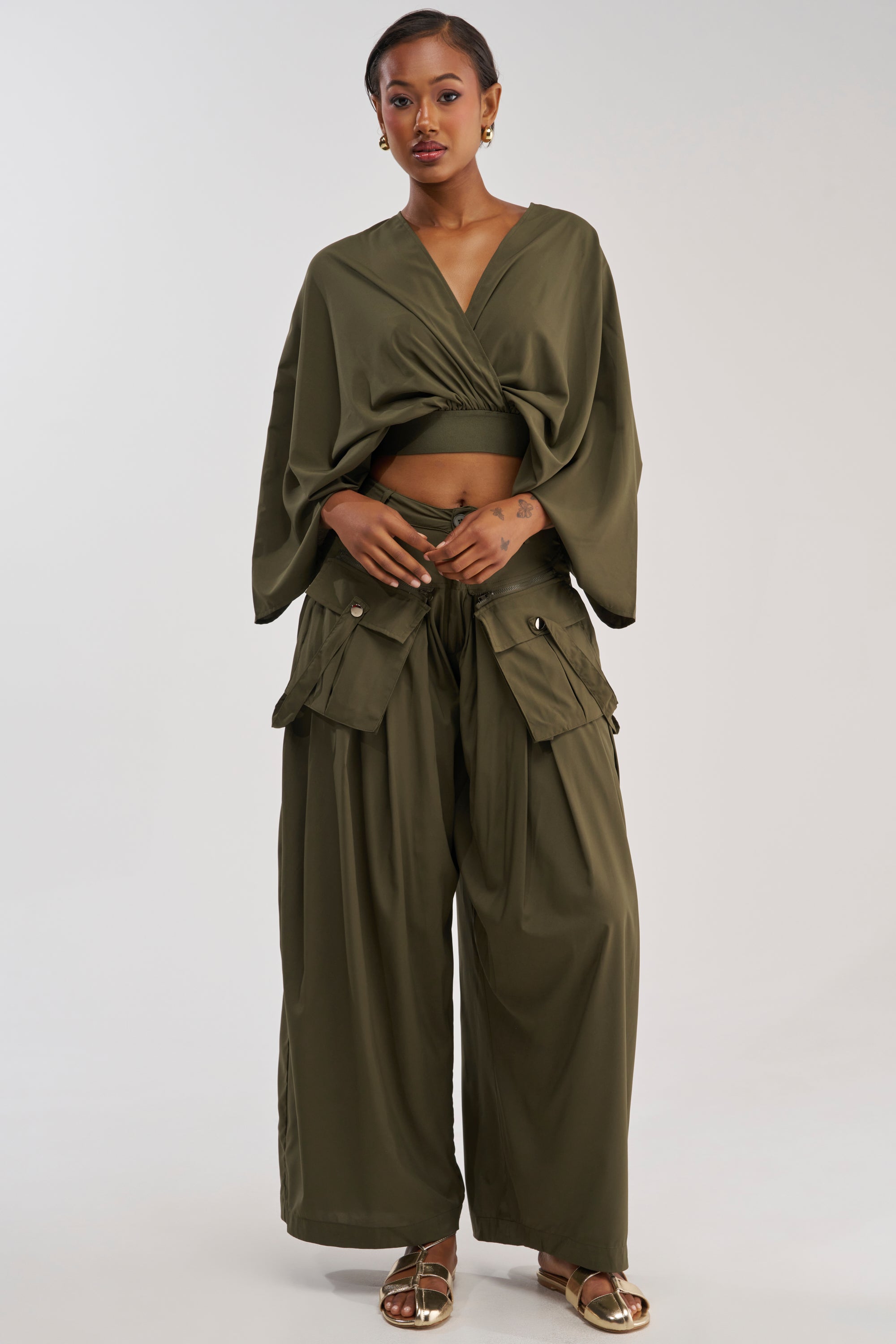 A woman models the TALK ABOUT ME TROUSER, pairing the utility-chic olive green pants with a matching long-sleeve v-neck crop top and gold sandals, set against a plain light background.