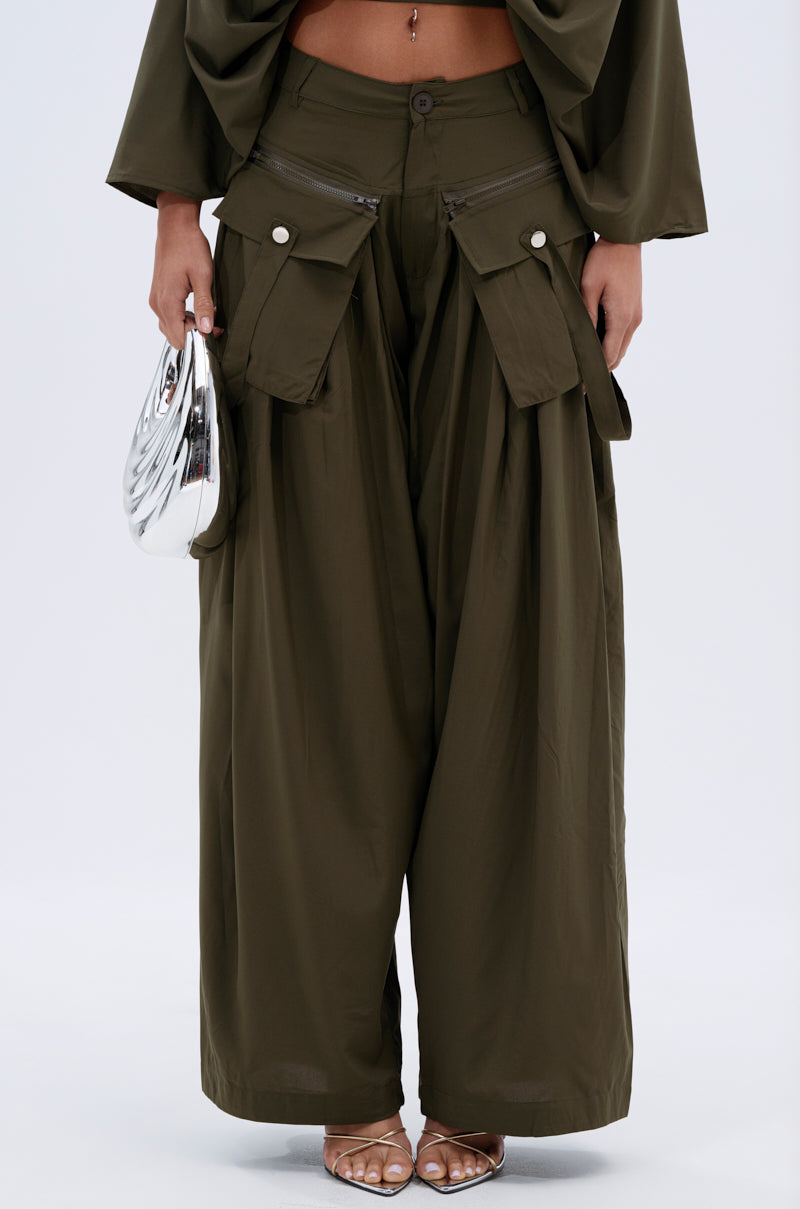 A person wearing the AKIRA TALK ABOUT ME TROUSER in olive green with pleats, large pockets, and zippers, styled with a small metallic silver clutch and heeled sandals. 1