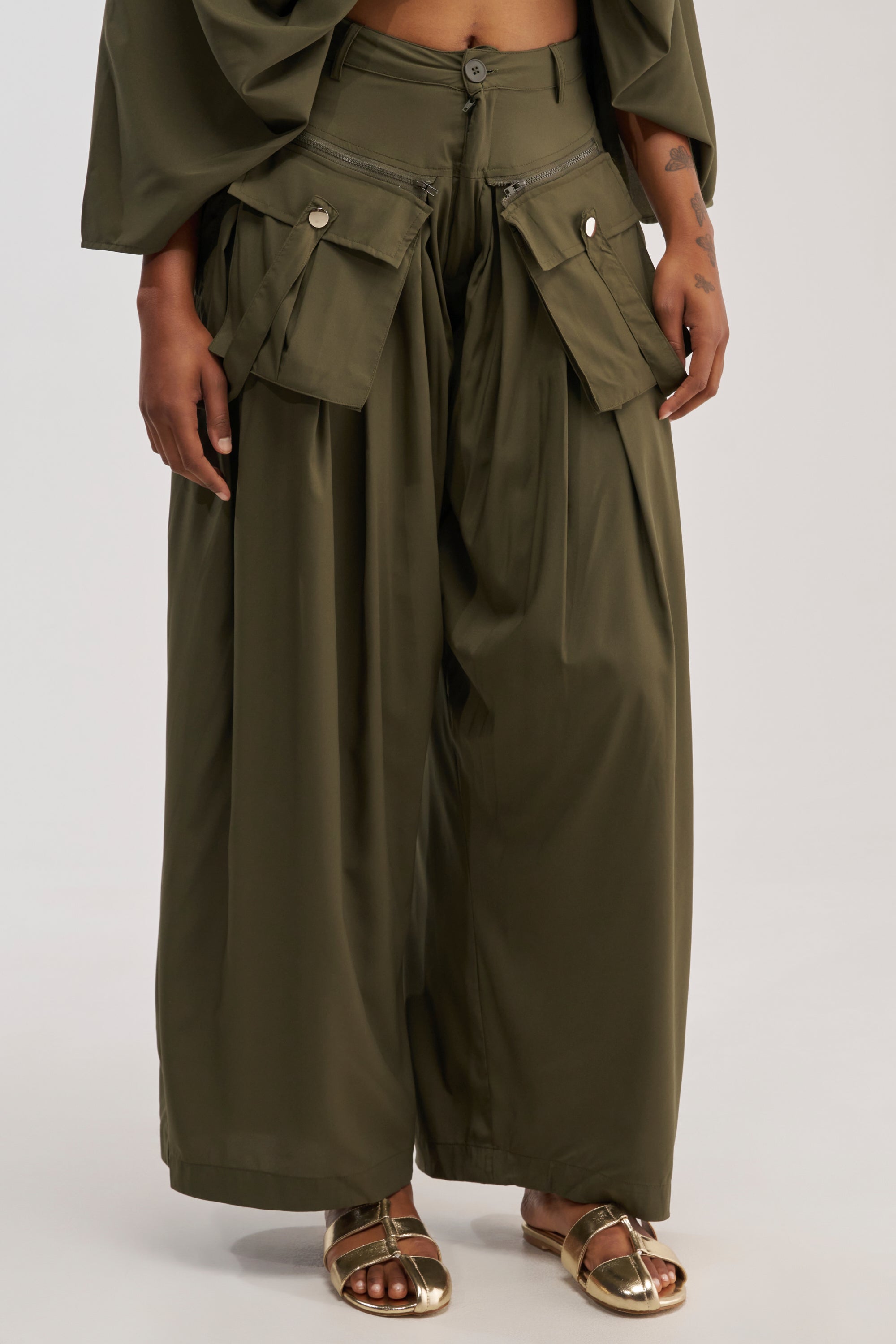 A person is shown wearing the TALK ABOUT ME TROUSER, styled in olive green with a wide-leg silhouette and large side pockets, paired with gold sandals. The lower half of their body is pictured against a plain background, highlighting the modern utility-chic design of the pants.