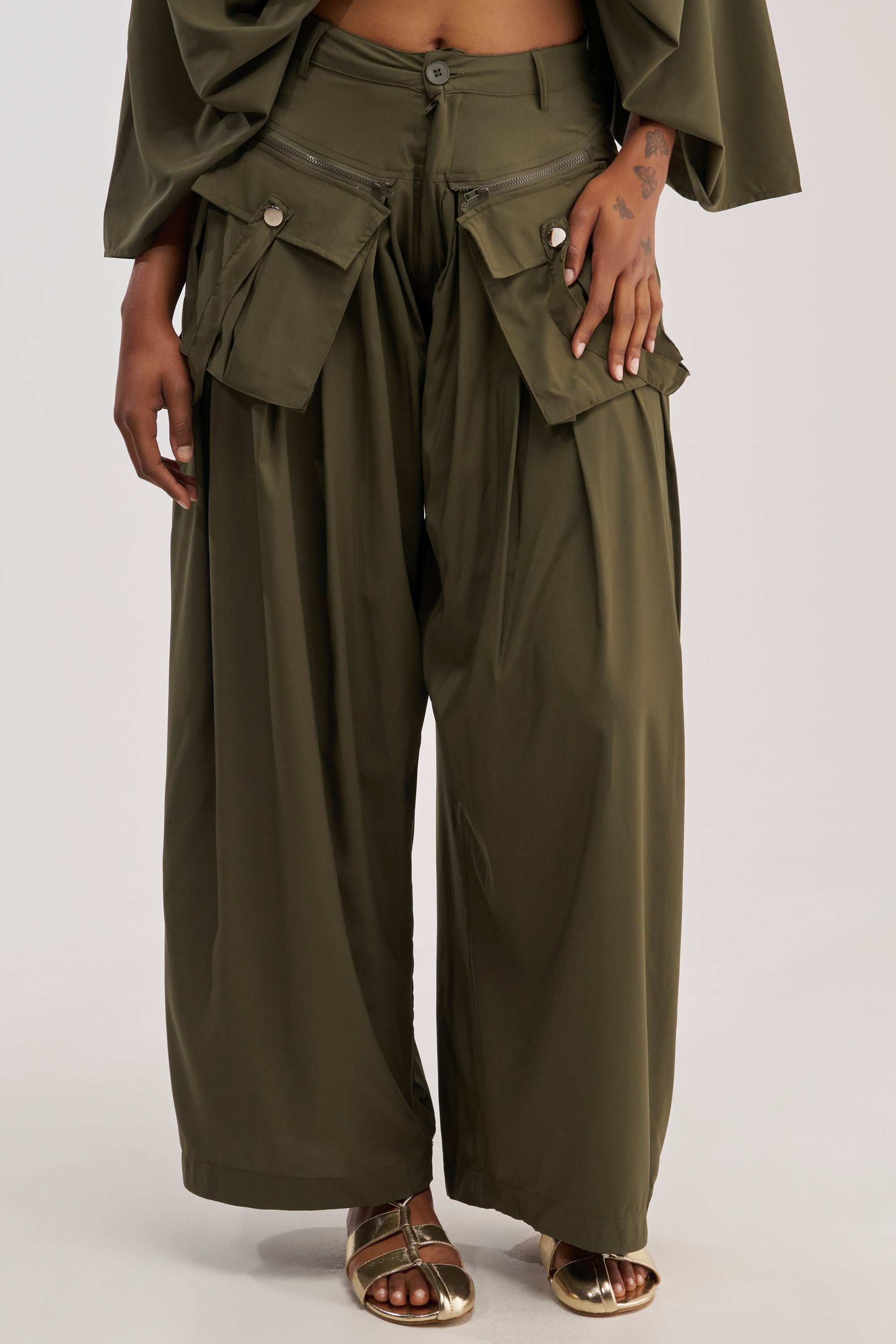 A person showcases the lower half of their outfit, donning the TALK ABOUT ME TROUSER in olive—wide-leg bottoms featuring large front flap pockets. Their hand rests casually on one pocket, accentuating the trouser's cargo-inspired, utility-chic vibe. The look is completed with a matching loose top and gold sandals.