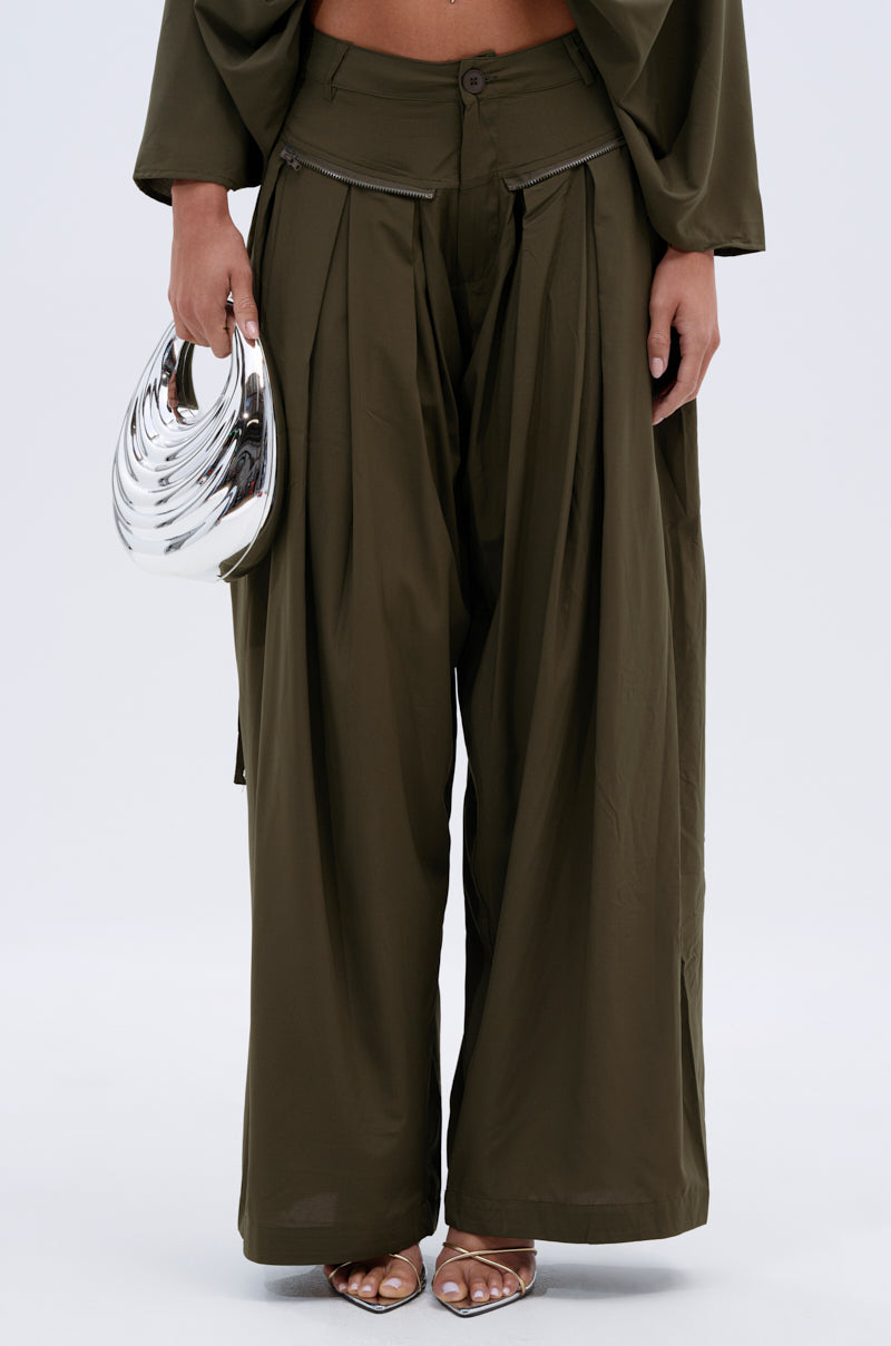 A person wears the TALK ABOUT ME TROUSER—loose-fitting, olive green, pleated cargo pants—paired with a matching top and silver crescent handbag. They sport open-toed heels in an image cropped at the waist and ankles. 3