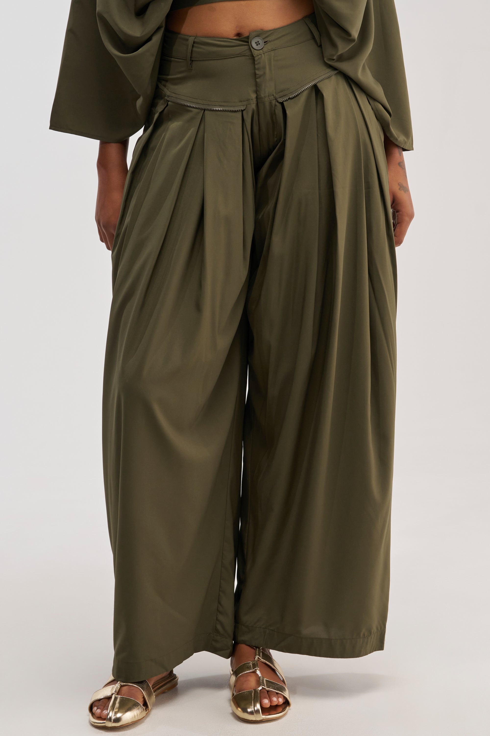 Wearing the TALK ABOUT ME TROUSER—olive wide-leg, high-waisted pants with a loose fit, visible pleats, and distinctive overlapping front detail—paired with gold sandals. The utility-chic trousers stand out against a plain light background.