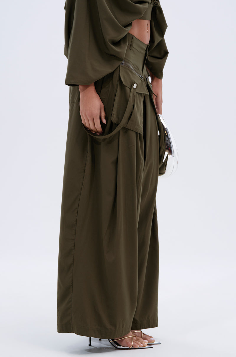 A person wears the TALK ABOUT ME TROUSER—AKIRA Label high-rise wide-leg pants with large front pockets—paired with heeled sandals and a metallic clutch, standing against a plain light background. 4